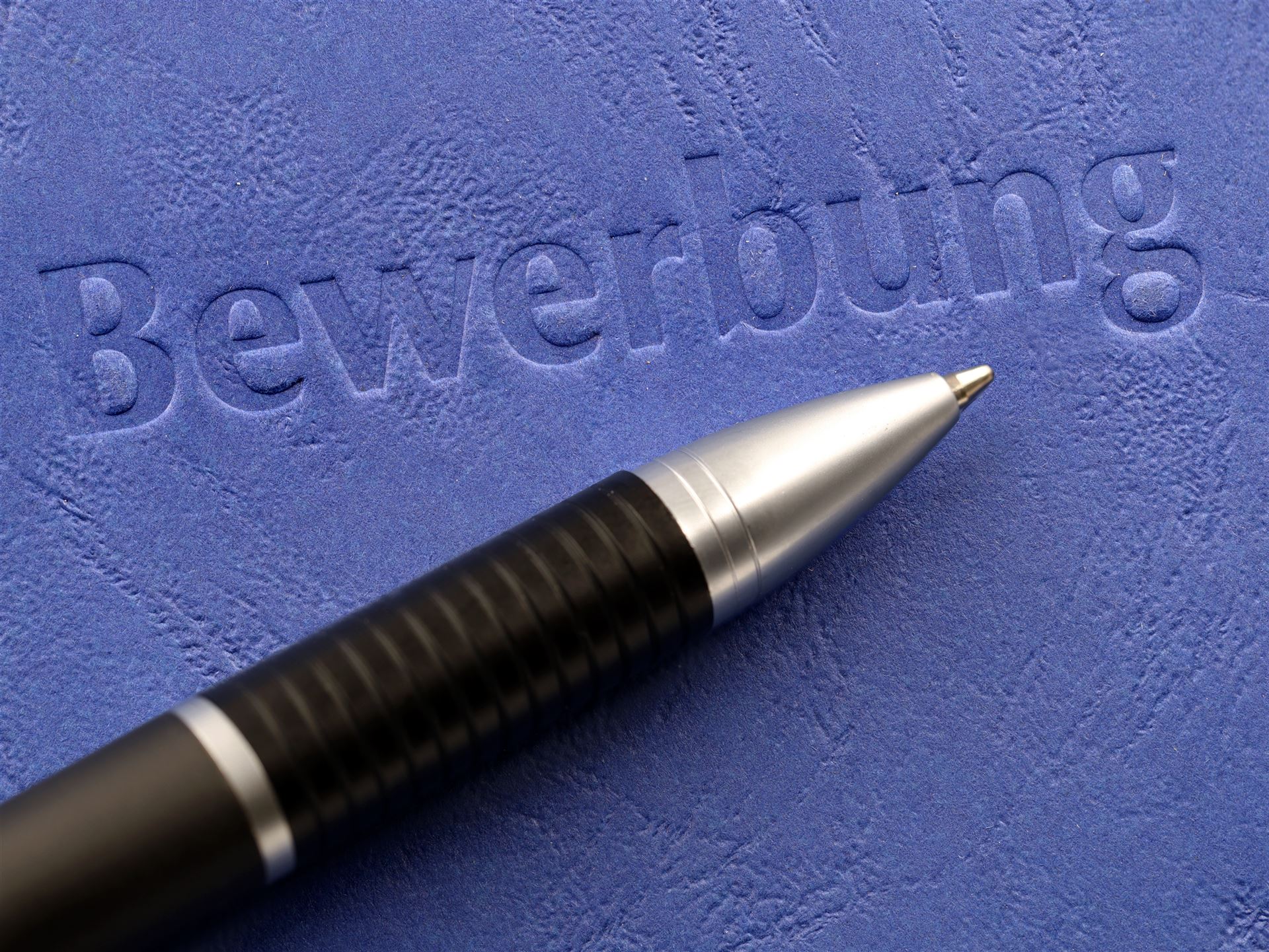 Bild zeigt auf blauem Hintergrund das eingeprägte Word "Bewerbung". Drunter liegt ein Kugelschreiber
