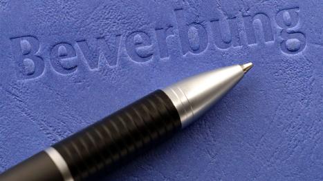 Bild zeigt auf blauem Hintergrund das eingeprägte Word "Bewerbung". Drunter liegt ein Kugelschreiber